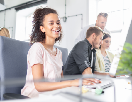 Glückliche Mitarbeiterin arbeitet am PC. Kollegen arbeiten im Hintergrund.